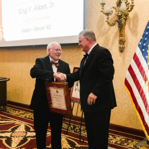 two men in tuxes shaking hands after one receives framed certificate