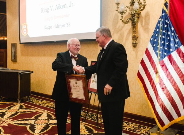 two men in tuxes shaking hands after one receives framed certificate