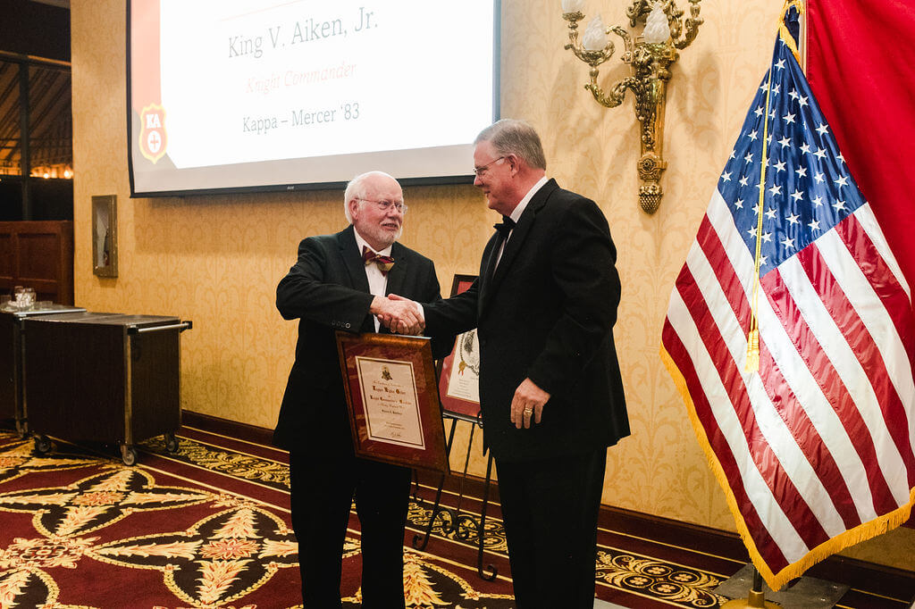 two men in tuxes shaking hands after one receives framed certificate