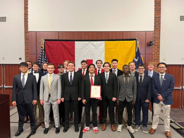 group of young men in suits in front of large KA flag holding certificate