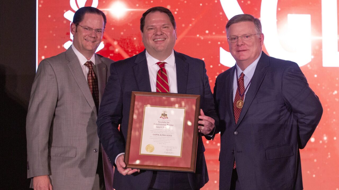 three men in suits with center holding framed certificate